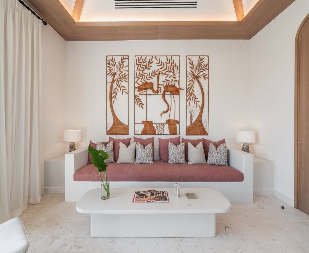 Sala de estar con sillón blanco, cojines decorativos y una mesa blanca con una planta, un libro abierto, y otros objetos. En la pared, arte de madera con diseño de flamencos y naturaleza. Lámparas a ambos lados de la sala y cortinas blancas a la izquierda.