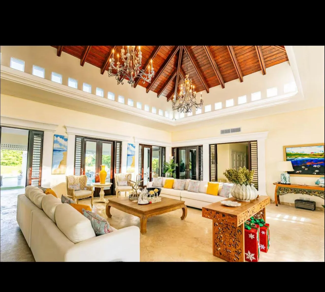 Sala de estar con techos de madera, candelabros y grandes ventanas que dan al exterior, muebles blancos y decoraciones coloridas.