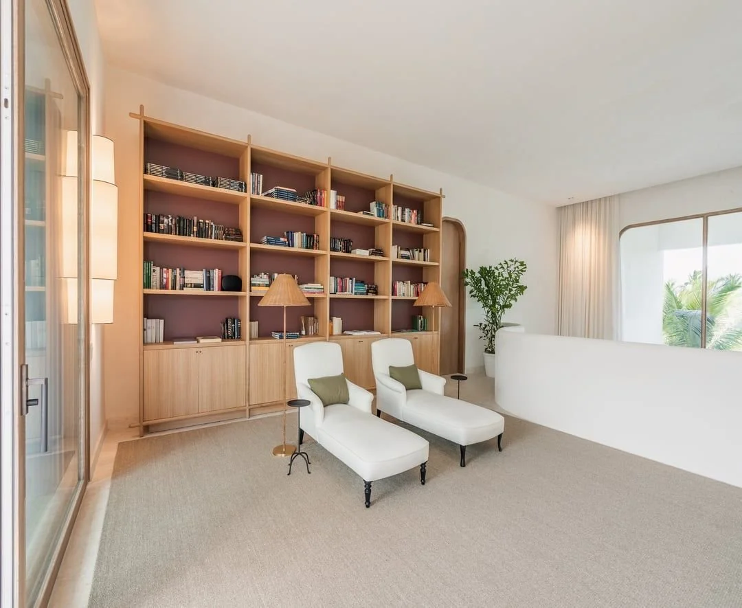 Sala con estantería de madera llena de libros, dos sillones blancos con cojines verdes, lámparas de mesa, planta y ventanas con cortinas blindadas, vista hacia árboles verdes.