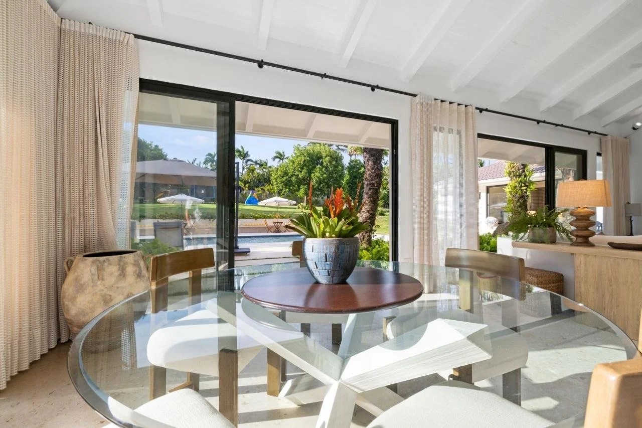 Comedor con mesa de vidrio, planta en jarrón en el centro, ventanas grandes con cortinas transparentes, vista a un jardín con alberca y áreas de descanso.