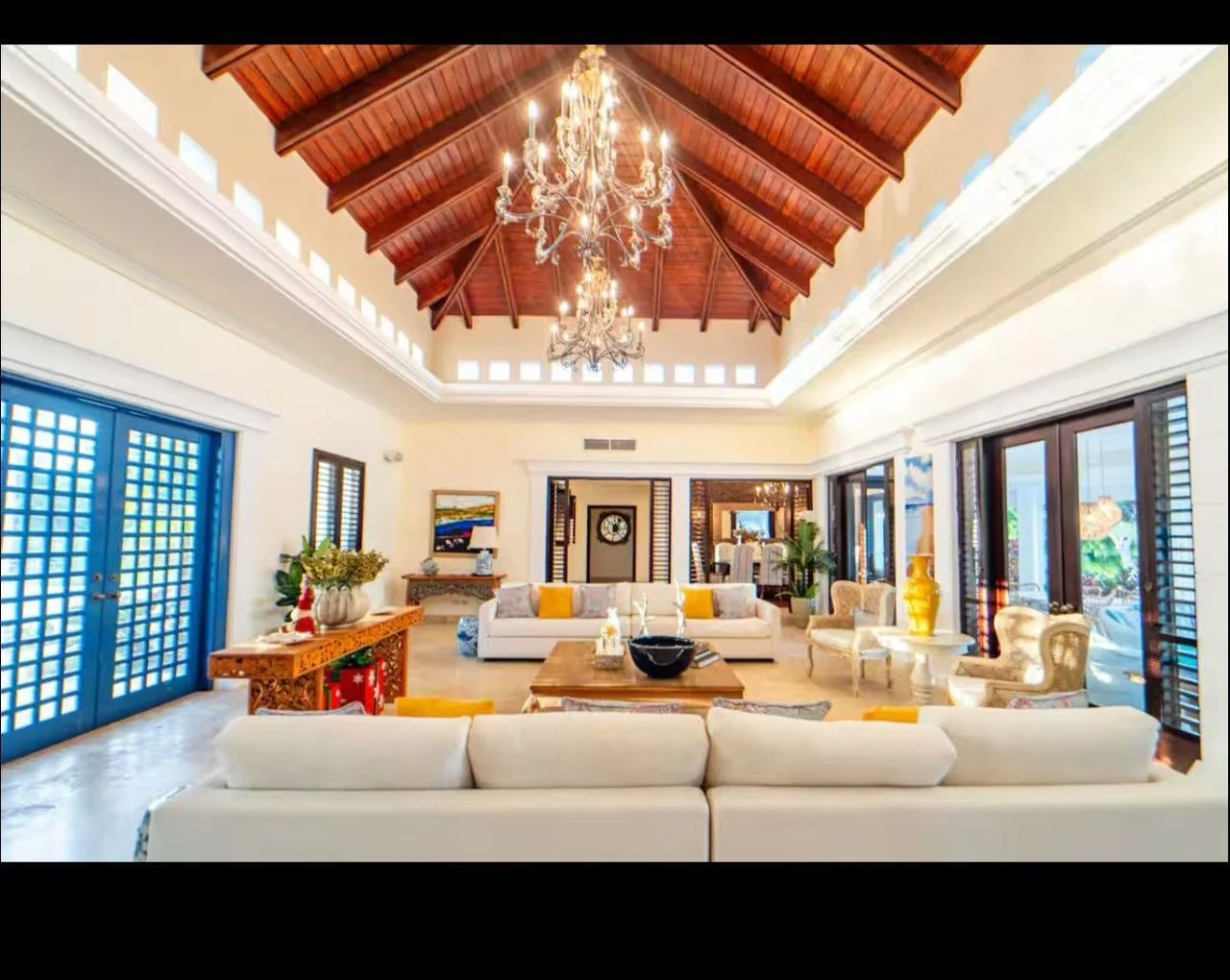 Sala de estar con techo alto de madera, candelabros, sillones blancos, ventanas grandes con persianas, mesa central de madera, decoración en colores claros y detalles dorados.