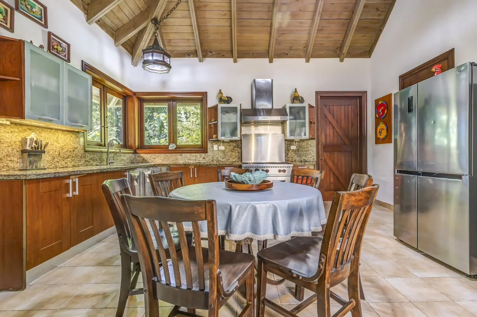 Cocina rústica con mesa redonda y siete sillas de madera, ventanas grandes, electrodomésticos de acero inoxidable y techo de madera