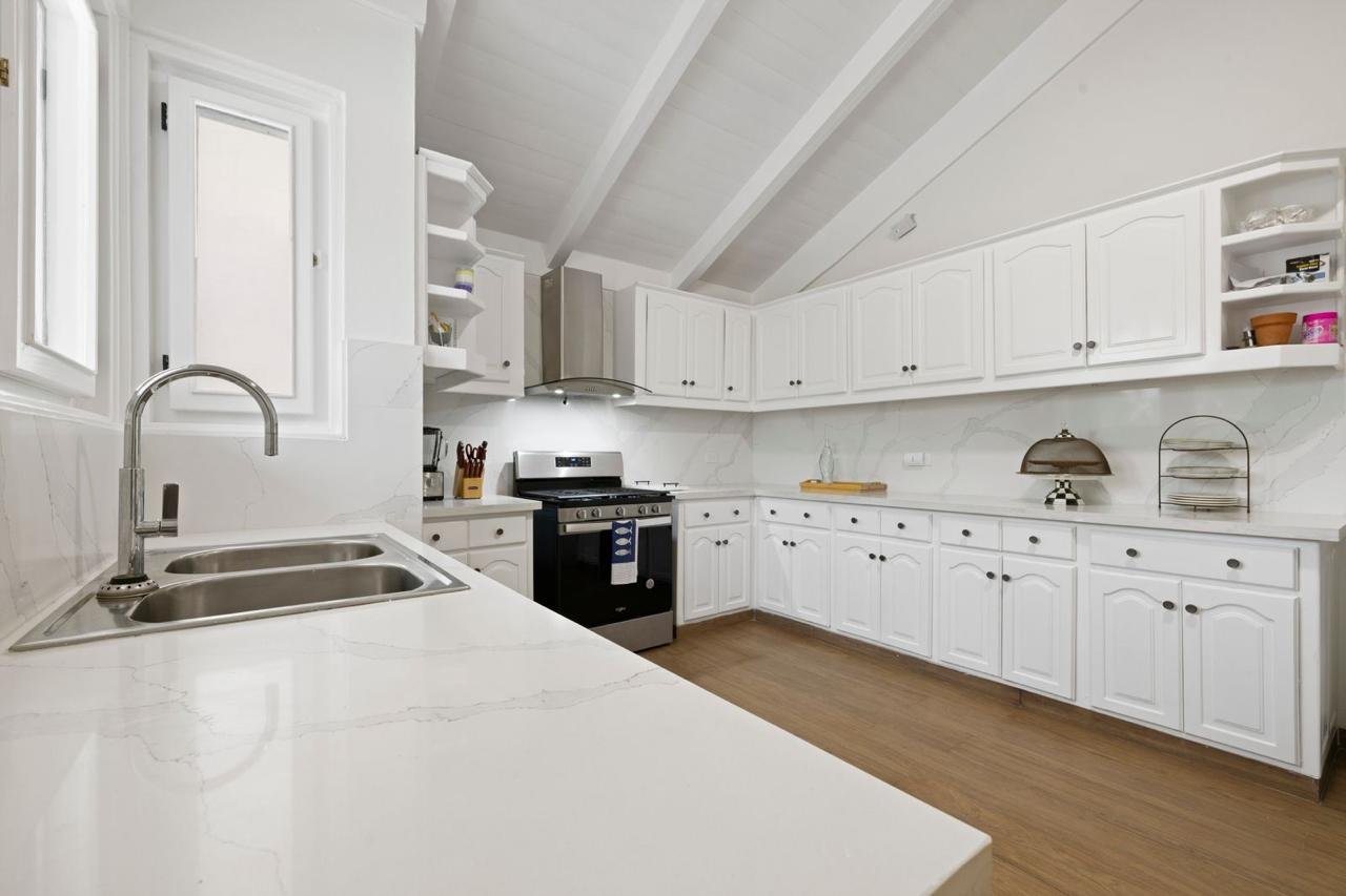 Cocina moderna con encimeras blancas y gabinetes blancos, estufa negra, fregadero doble de acero inoxidable, suelo de madera, ventanas y algunos objetos decorativos en las encimeras.