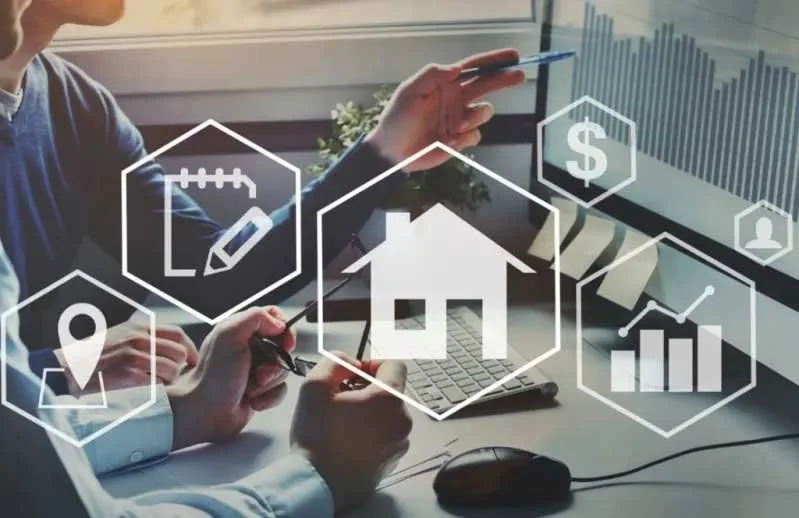 Personas trabajando en una mesa con íconos digitales de bienes raíces, finanzas y negocios, incluyendo una casa, una gráfica y un dólar.