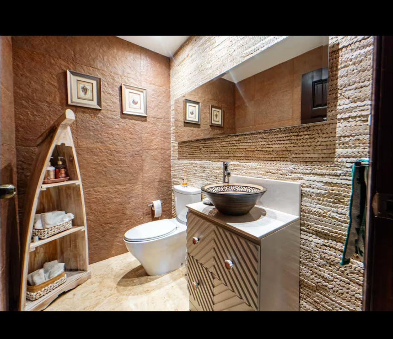 Baño con paredes de piedra y decoracion natural, lavabo en mueble de madera rayada, espejo grande, inodoro, estante en forma de barco con toallas y productos, tres cuadros con mariposas, y accesorios de baño.