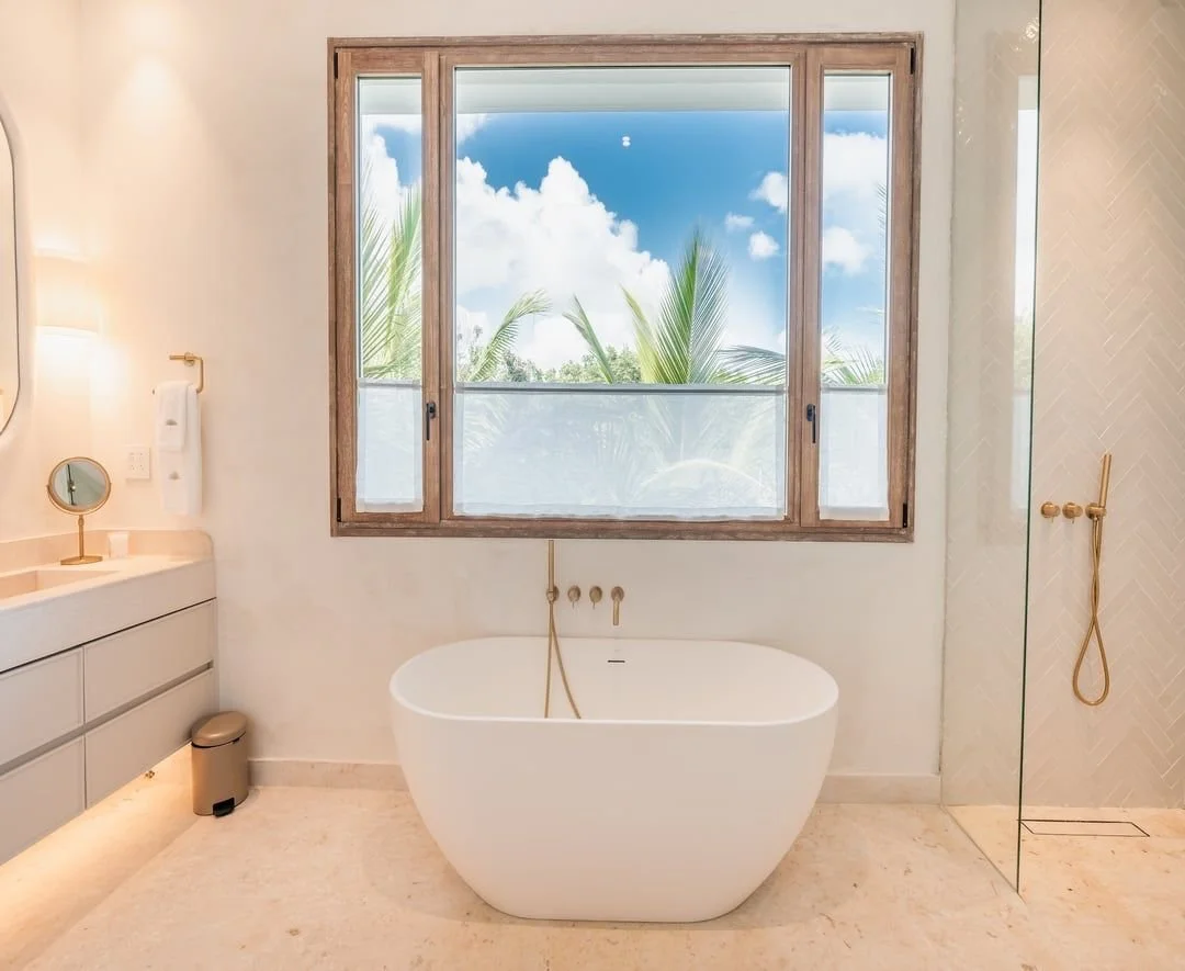 Cuarto de baño con bañera frente a una ventana grande, ducha a la derecha, lavabo con espejo a la izquierda, y vista de palmas y cielo azul con nubes.
