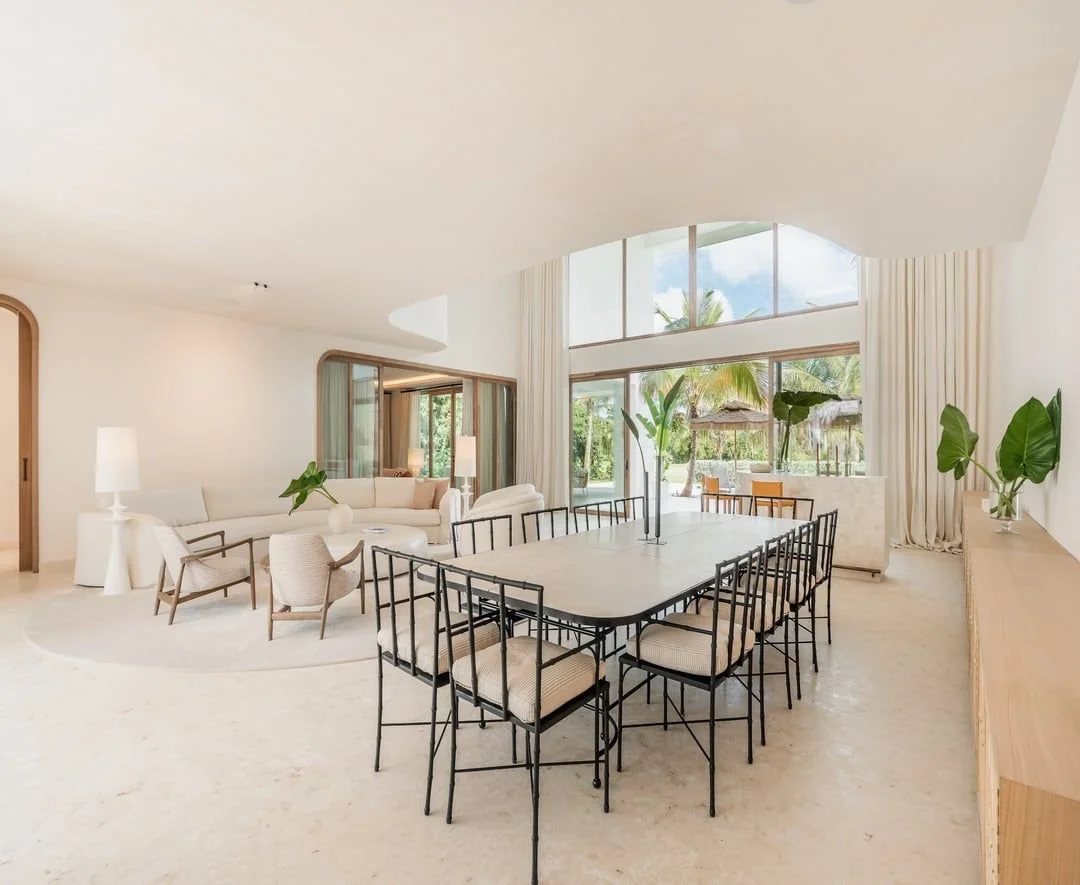 Sala de estar y comedor con muebles blancos y detalles de plantas verdes, con grandes ventanas que dejan entrar luz natural y muestran un exterior con palmeras.