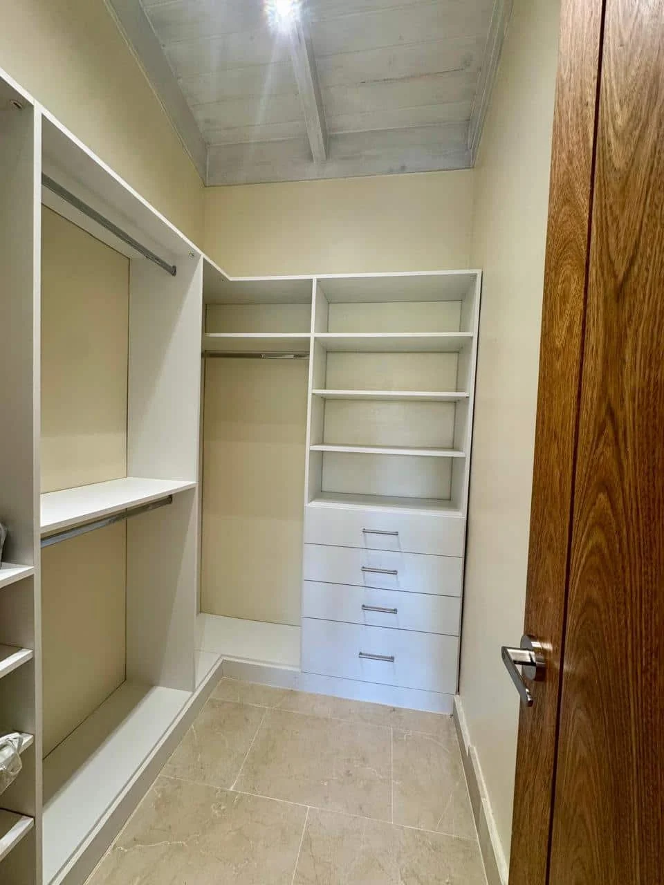 Armario de closet con estantes, cajones y barra para colgar ropa, en una habitación con piso de baldosas y puerta de madera.