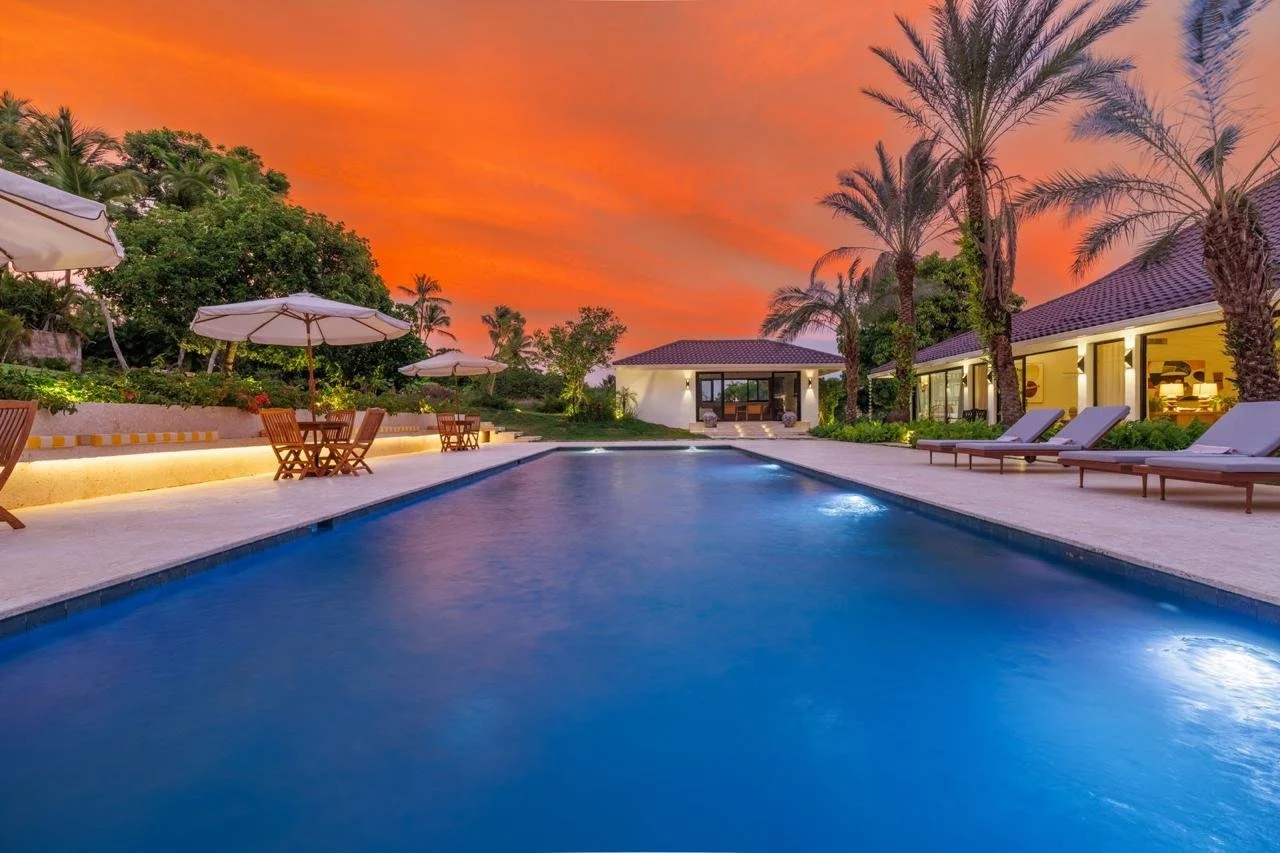 Piscina en un complejo residencial al atardecer con sombrillas, sillas reclinables, palmeras y una casa moderna con ventanas grandes.
