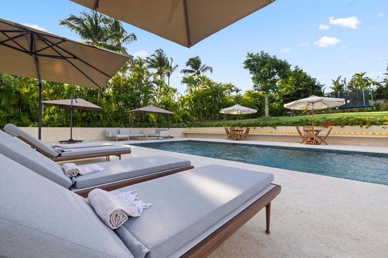 Cadeiras reclinables con toallas a la orilla de una piscina en un entorno tropical con árboles y sombrillas.