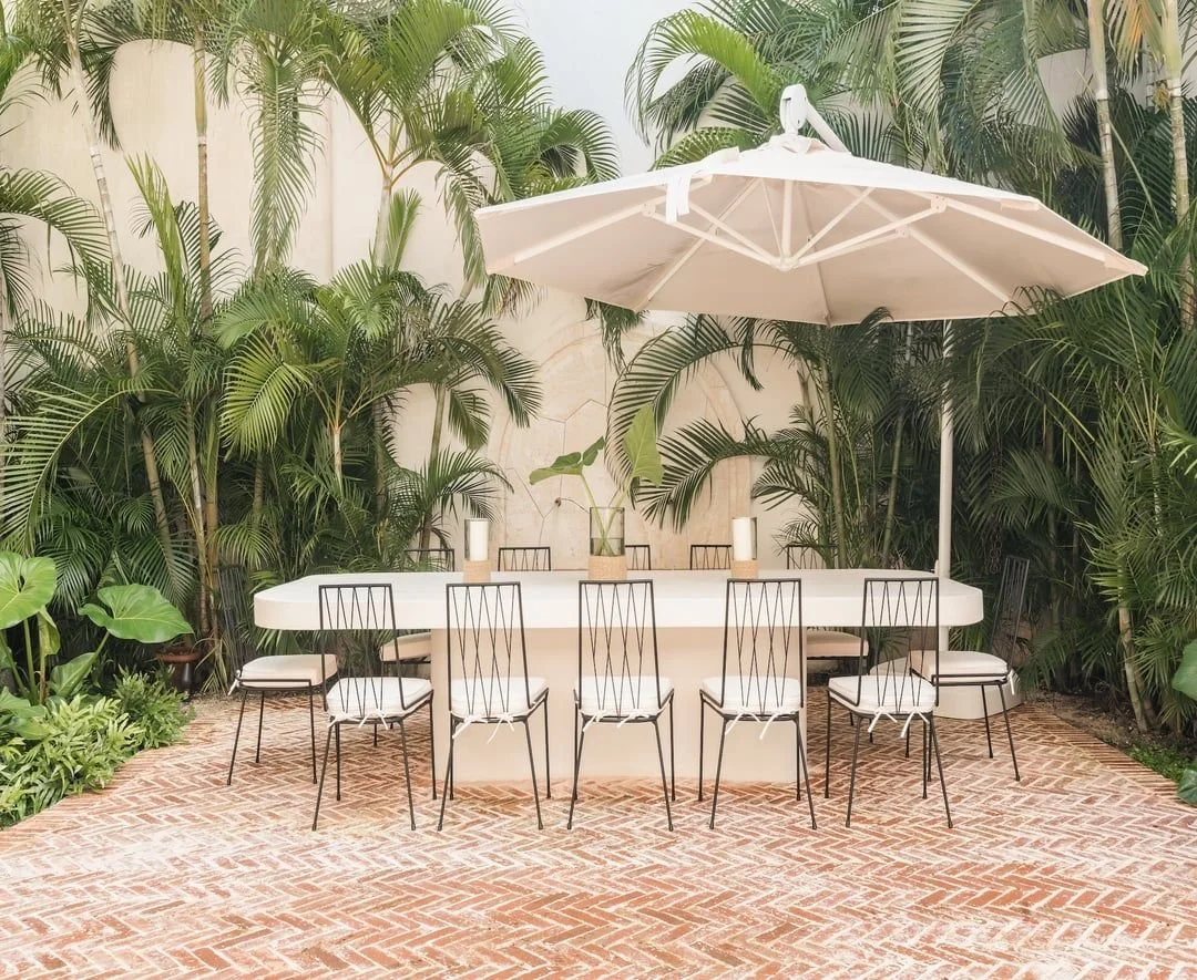 mesa blanca semicircular con siete sillas de metal negro y cojines blancos, toldo blanco, rodeada de plantas verdes tropicales, en patio con piso de ladrillos.