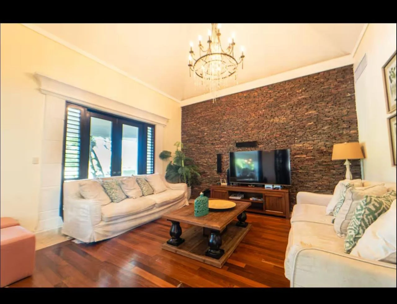 Sala de estar con dos sofás blancos, mesa de madera, televisor y pared de ladrillos en el fondo, lámpara de pie y varias plantas.