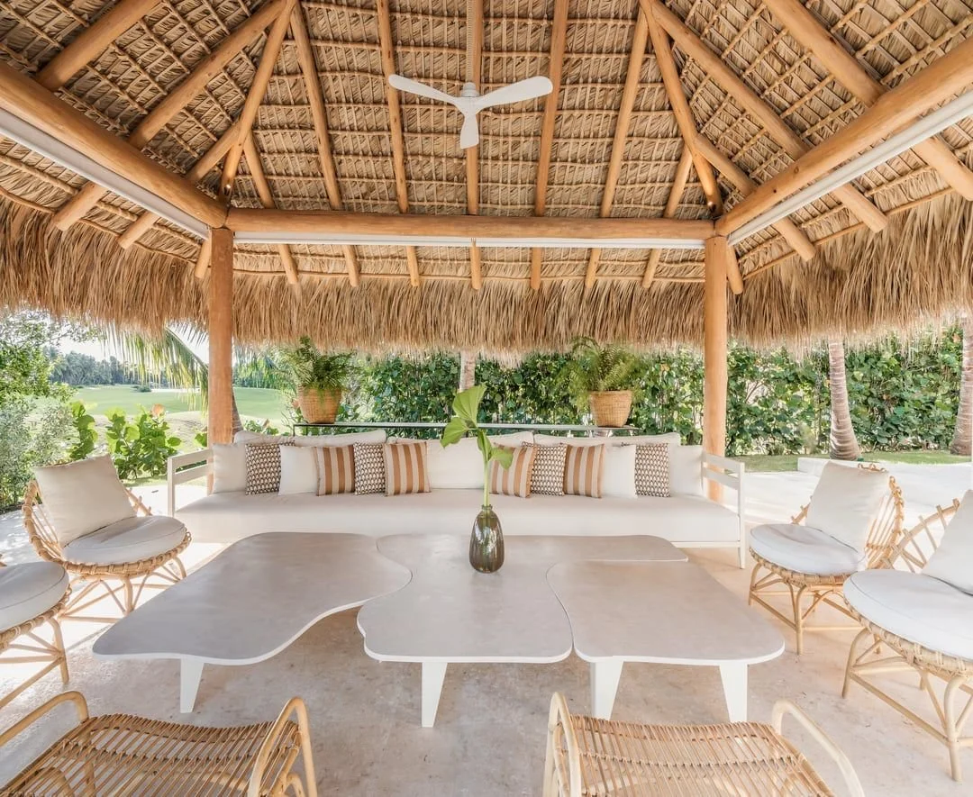 Una sala de estar al aire libre con muebles de mimbre y cojines blancos, bajo un techo de paja con ventilador de hélice, rodeada de vegetación y vista a un jardín.