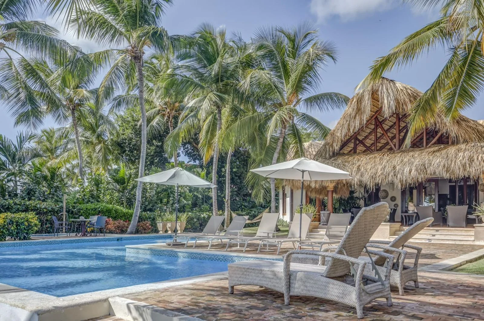 Piscina en un resort con sillas y sombrillas, rodeada de palmeras y un edificio con techo de paja.