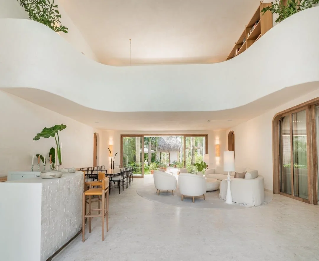 Sala de estar luminosa y moderna con muebles blancos y grandes ventanas que muestran un jardín tropical