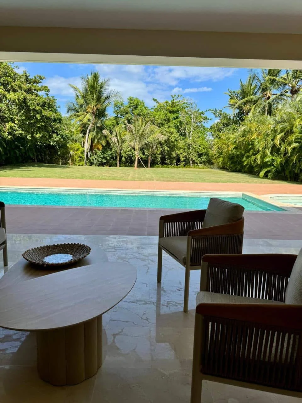 Vista de una piscina con césped y árboles tropicales, vista desde un patio con sillas de madera y una mesa redonda en un entorno de clima cálido.