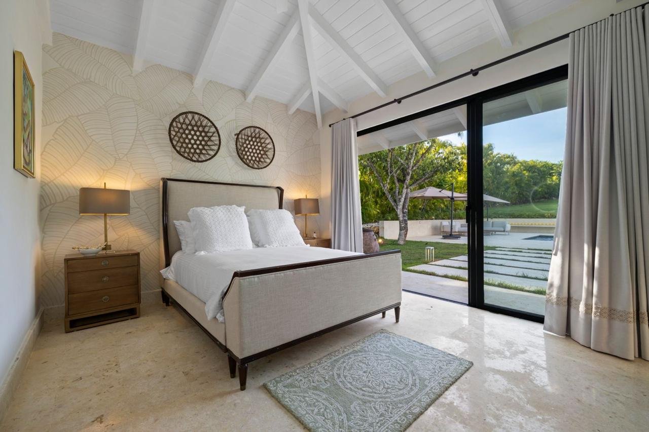 Habitación con cama grande, dos mesitas de noche con lámparas, cortinas blancas, vista al jardín con árboles y sombra, y decoración moderna.