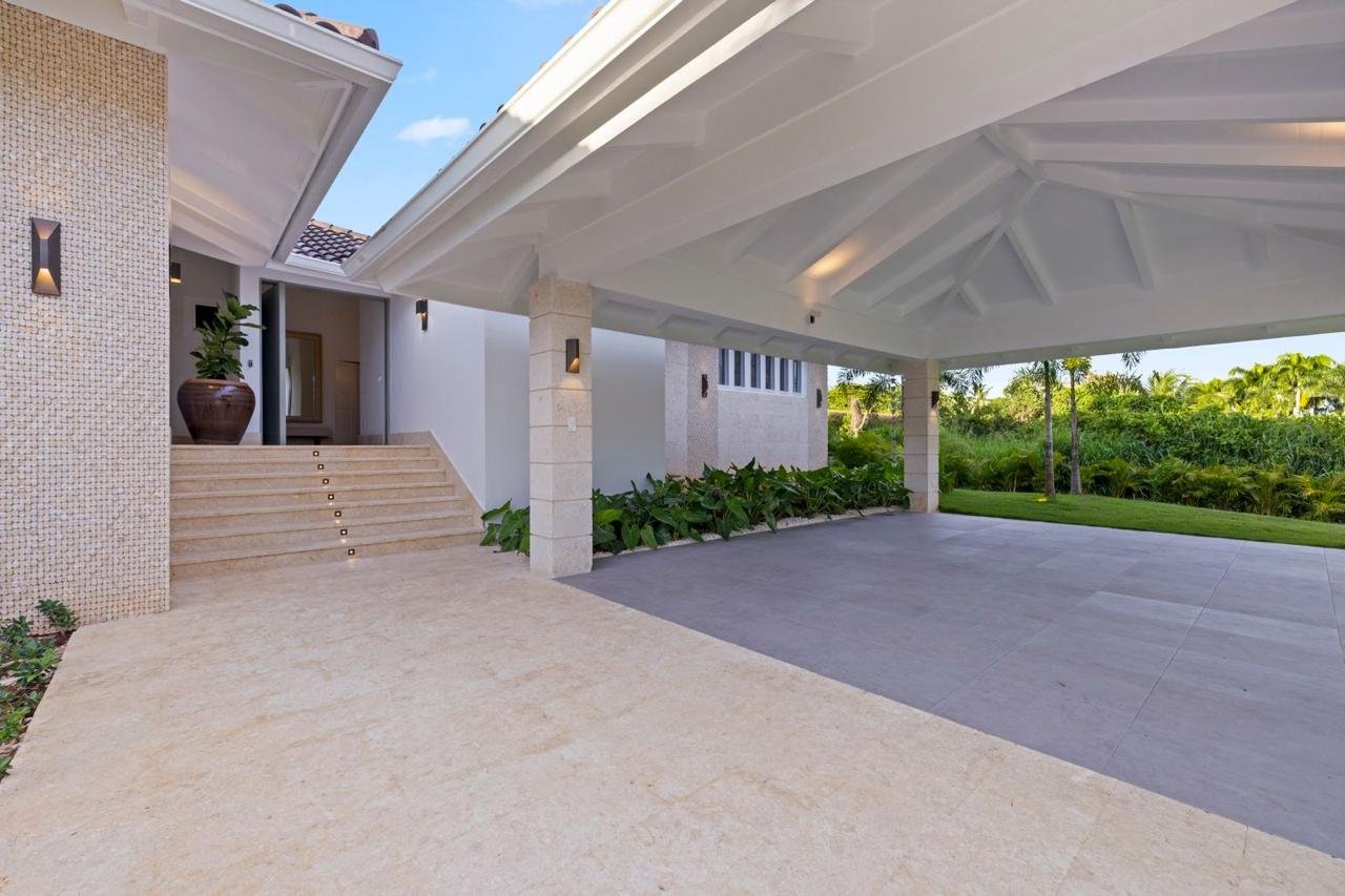 Exterior de una casa moderna con entrada y área techada, rodeada de vegetación tropical y un cielo despejado.
