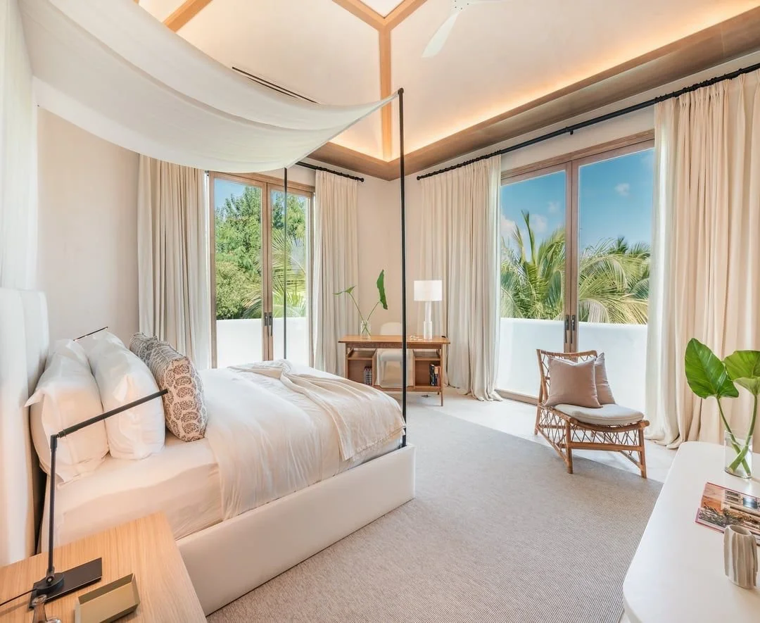 Habitación moderna con cama king size, cortinas blancas, gran vista a árboles y palmeras, sillón de mimbre, lámpara y escritorio.