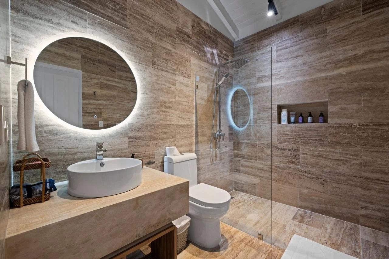 Baño moderno con paredes y pisos de piedra en tonos beige, lavabo ovalado sobre encimera de piedra, espejo redondo con luz LED, inodoro, ducha con techo de lluvia, pared con estantería de productos de baño, toalla colgada, y accesorios decorativos.