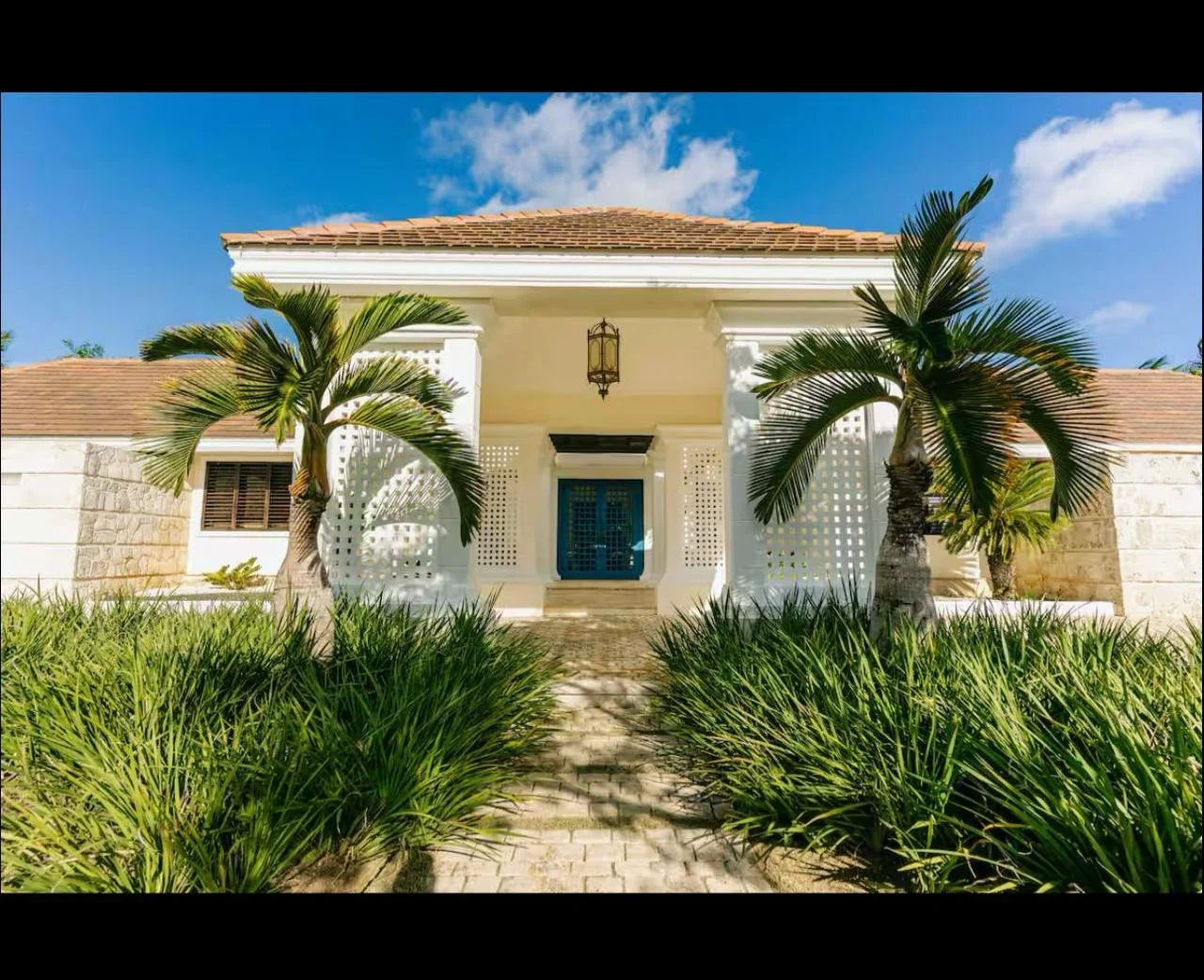Casa moderna con entrada azul, rodeada de palmeras y vegetación, bajo cielo soleado con nubes.