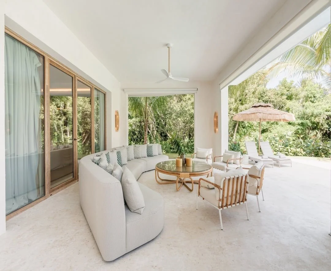 Salón con sofá blanco, sillas y mesa de centro en una terraza con vista a vegetación y playa, paraguas de paja y tumbonas.