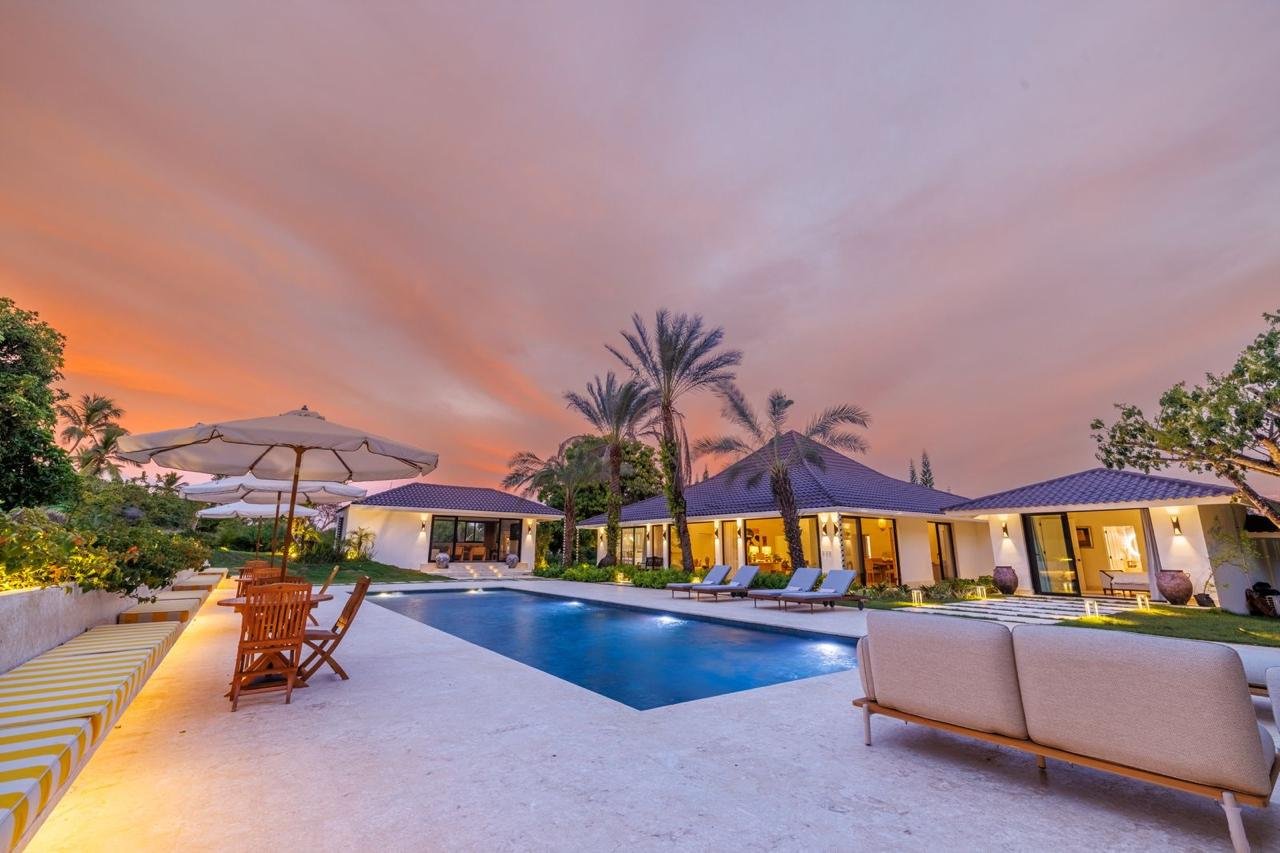 Piscina en el patio de una casa moderna con sillones, sillas de madera y sombrillas bajo un cielo color pastel