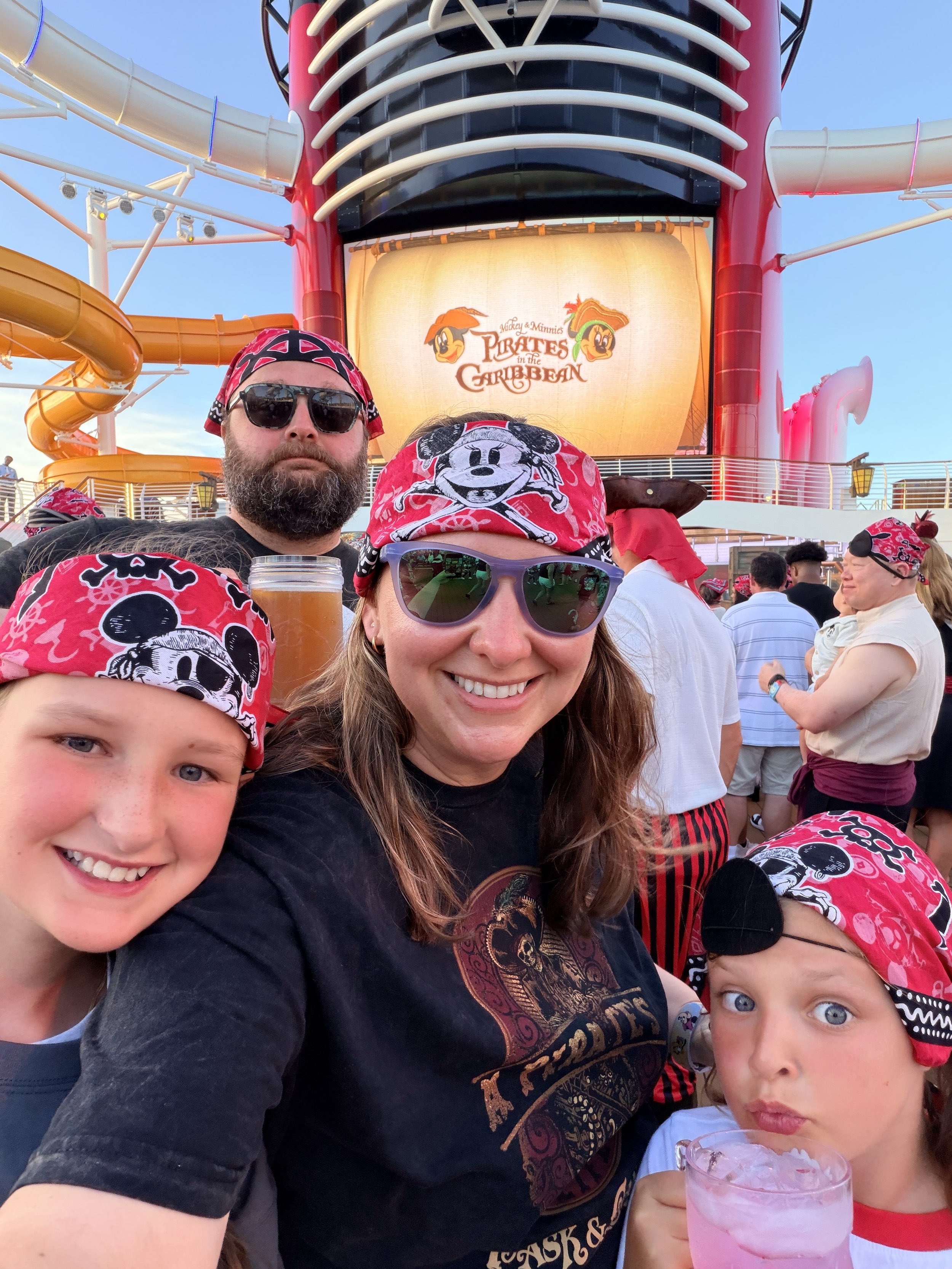 A group of people dressed as pirates on a cruise ship, with the ship's sign 'Mickey & Minnie's Pirates in the Caribbean' visible in the background.