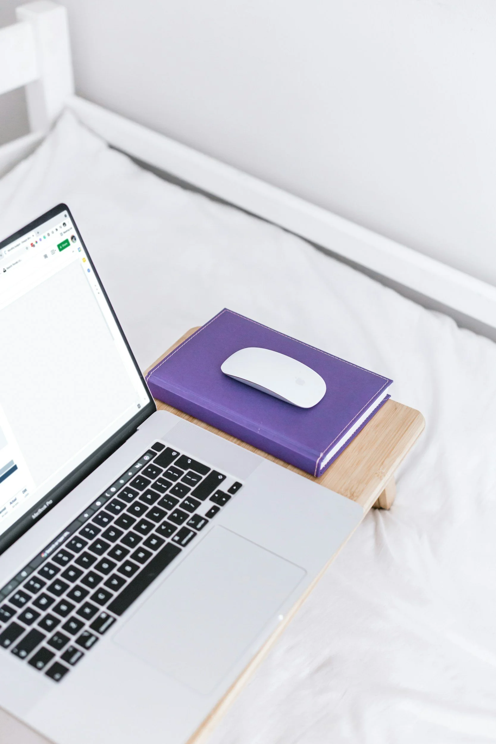 A laptop on a wooden desk with a white computer mouse on a purple notebook, in a bright white room.