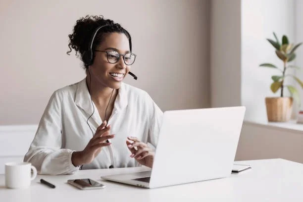 A woman smiling and wearing a headset, sitting at a desk with a laptop, in a well-lit room with a potted plant on the windowsill.