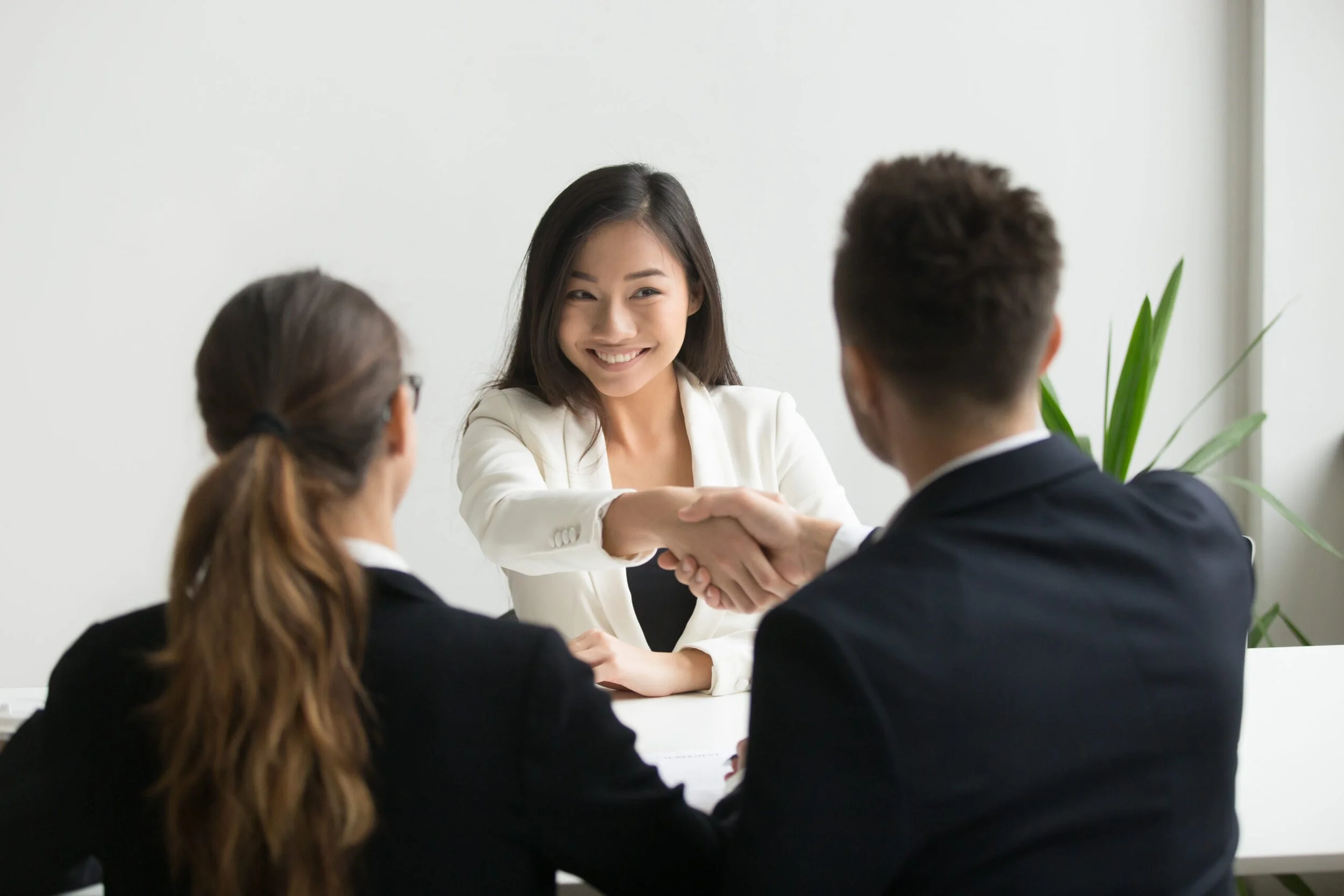 A woman in a white blazer shaking hands with a man in a suit during an interview in a modern office setting, with two women observing.