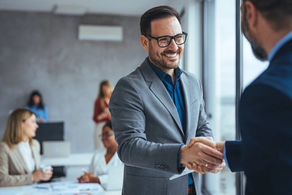 Two men shaking hands in a business meeting, with one man smiling and wearing glasses, inside a modern office.