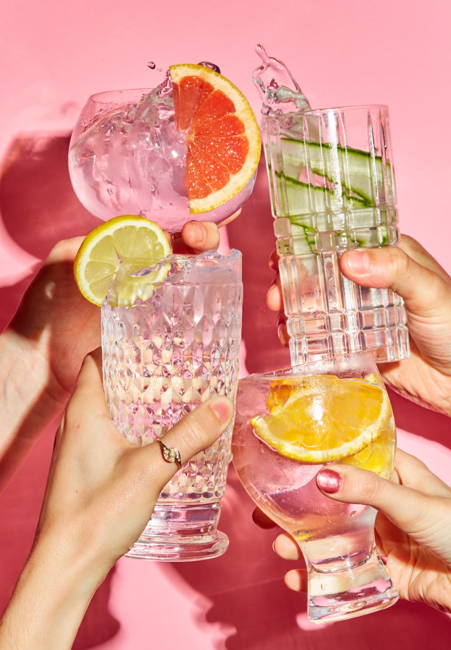 Four hands holding different glasses with colorful fruit garnishes, toasting against a pink background.
