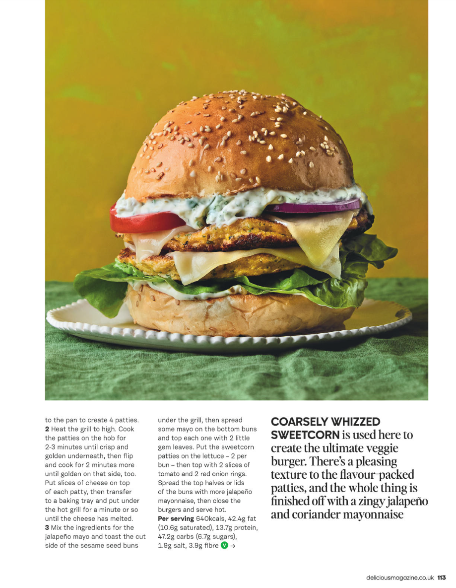 A large veggie burger with a sesame seed bun, lettuce, tomato, cheese, grilled veggie patty, onion, and a creamy sauce, served on a plate with a green cloth.