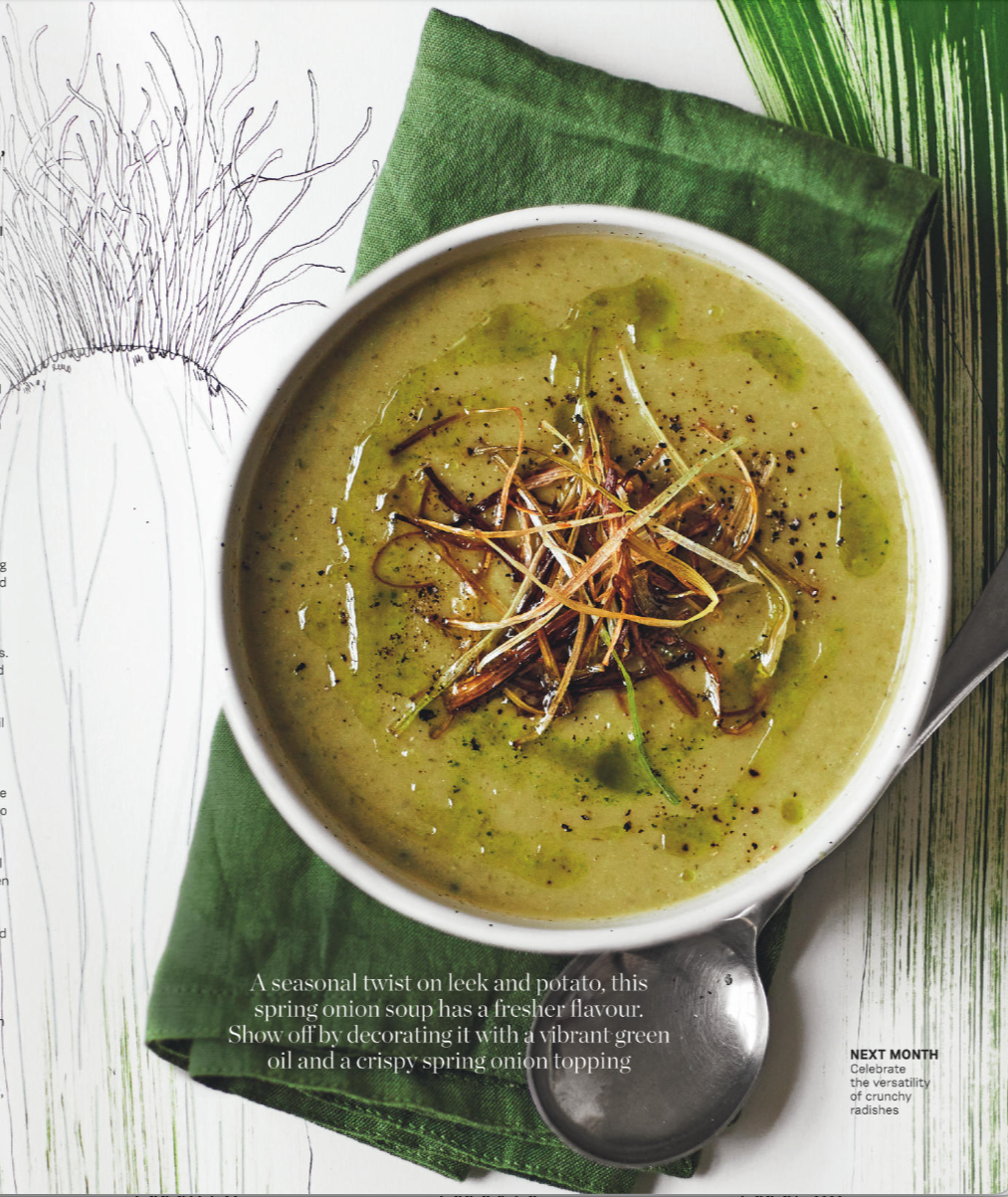 A bowl of spring onion soup topped with crispy spring onion slices and green oil. The bowl is placed on a green cloth napkin with a wooden table background.
