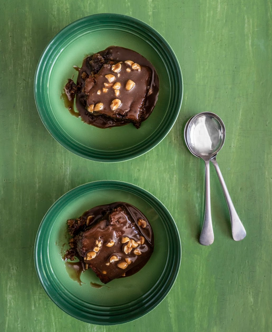 Two slices of chocolate cake with nuts in green bowls on a green wooden table, with a metal spoon beside them.