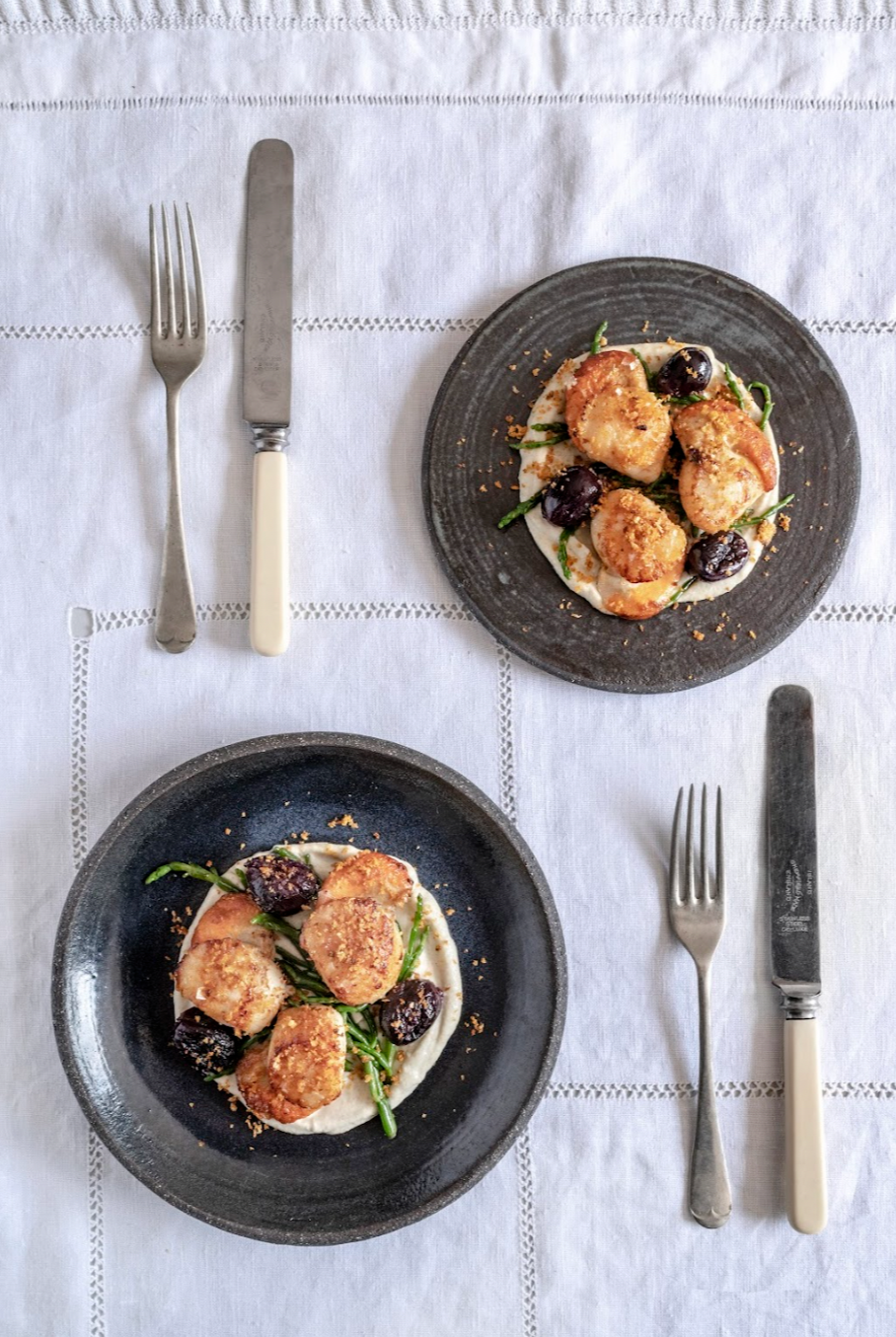 Two black bowls with cooked chicken breasts, dark purple vegetables, and green beans on white sauce, placed on a white tablecloth with cutlery.