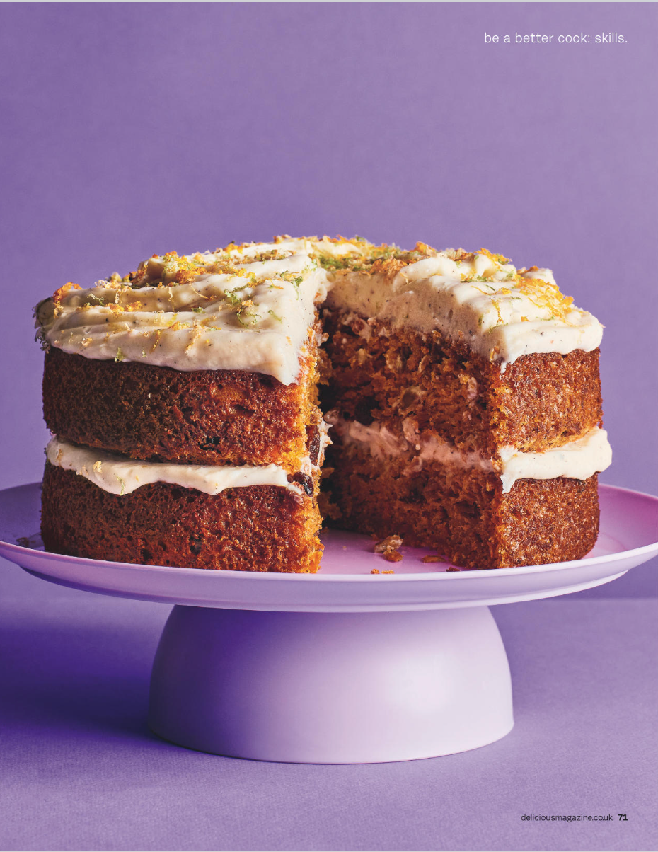 A layered carrot cake with cream cheese frosting and lemon zest on top, displayed on a pink cake stand against a purple background.