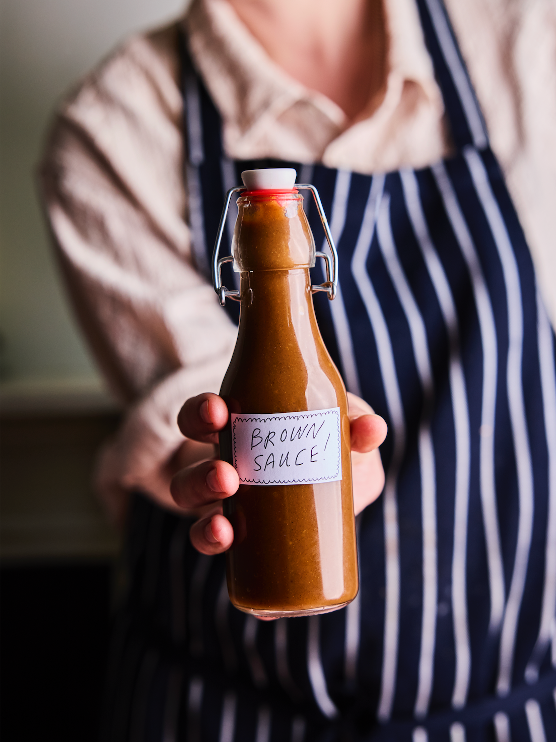 Person holding a glass bottle of brown sauce with a label that says 'Brown Sauce!' in handwritten text.