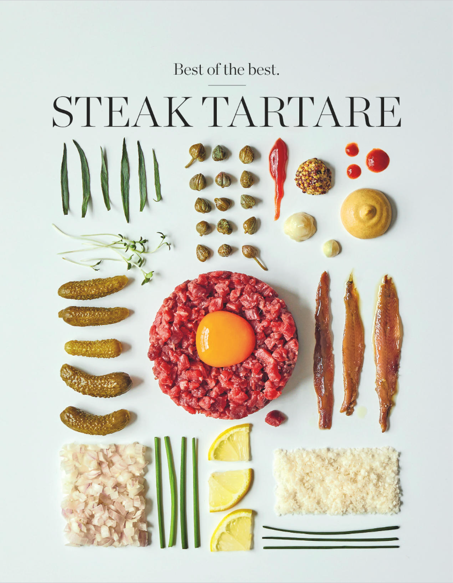 Arrangement of ingredients for a steak tartare dish, including raw beef, a raw egg yolk, capers, chopped onions, lemon wedges, and various sauces and garnishes, on a white background.