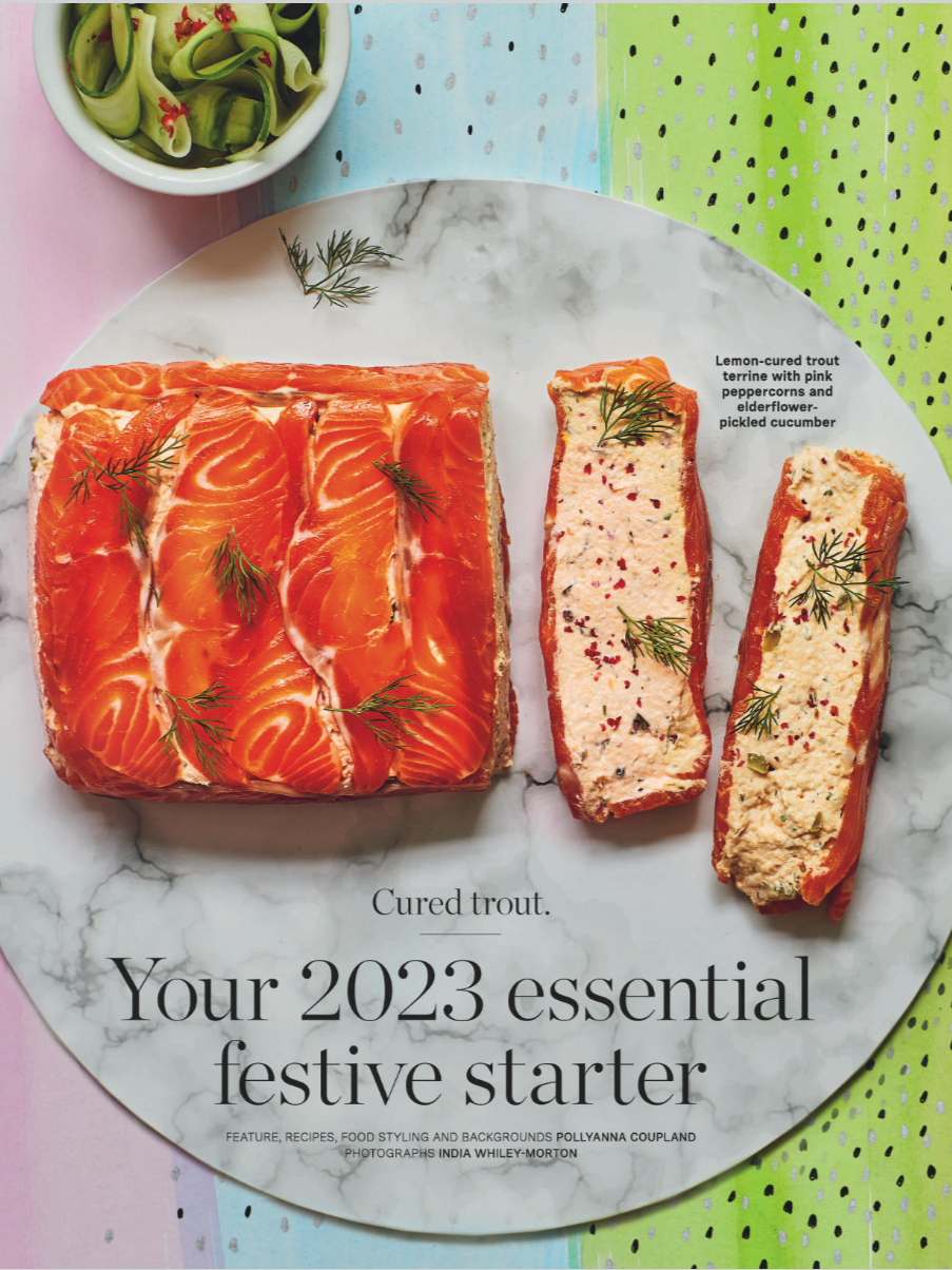A white marble platter with slices of cured trout topped with fresh dill, arranged artistically. A small bowl of cucumber salad with red pepper flakes is in the top left corner. The background features colorful pastel-colored paper with dotted patter