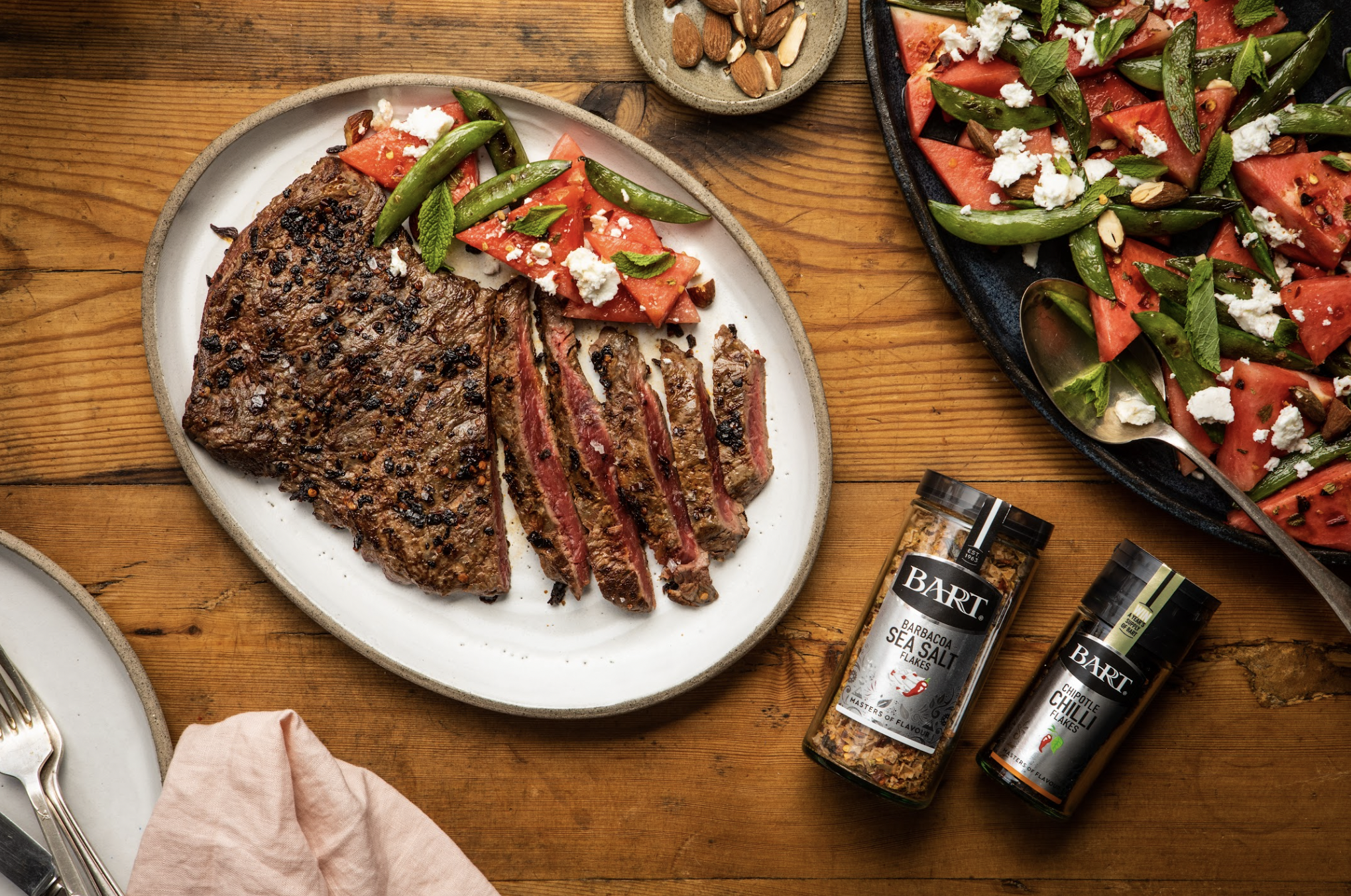 A wooden table with a sliced grilled steak topped with seasoning, served with a side of fresh tomato and green beans salad with crumbled cheese and mint leaves, and a bowl of mixed nuts.