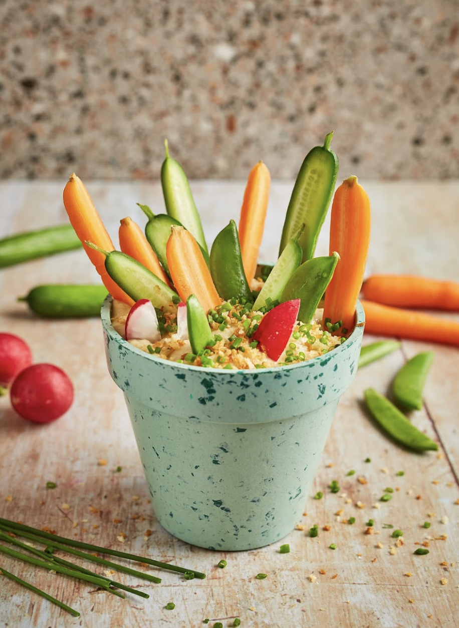 A blue speckled pot with colorful vegetable sticks including carrots, cucumbers, radishes, and snap peas, filled with hummus and garnished with chopped chives. Radish and vegetable pieces are scattered on a wooden surface with additional vegetables i