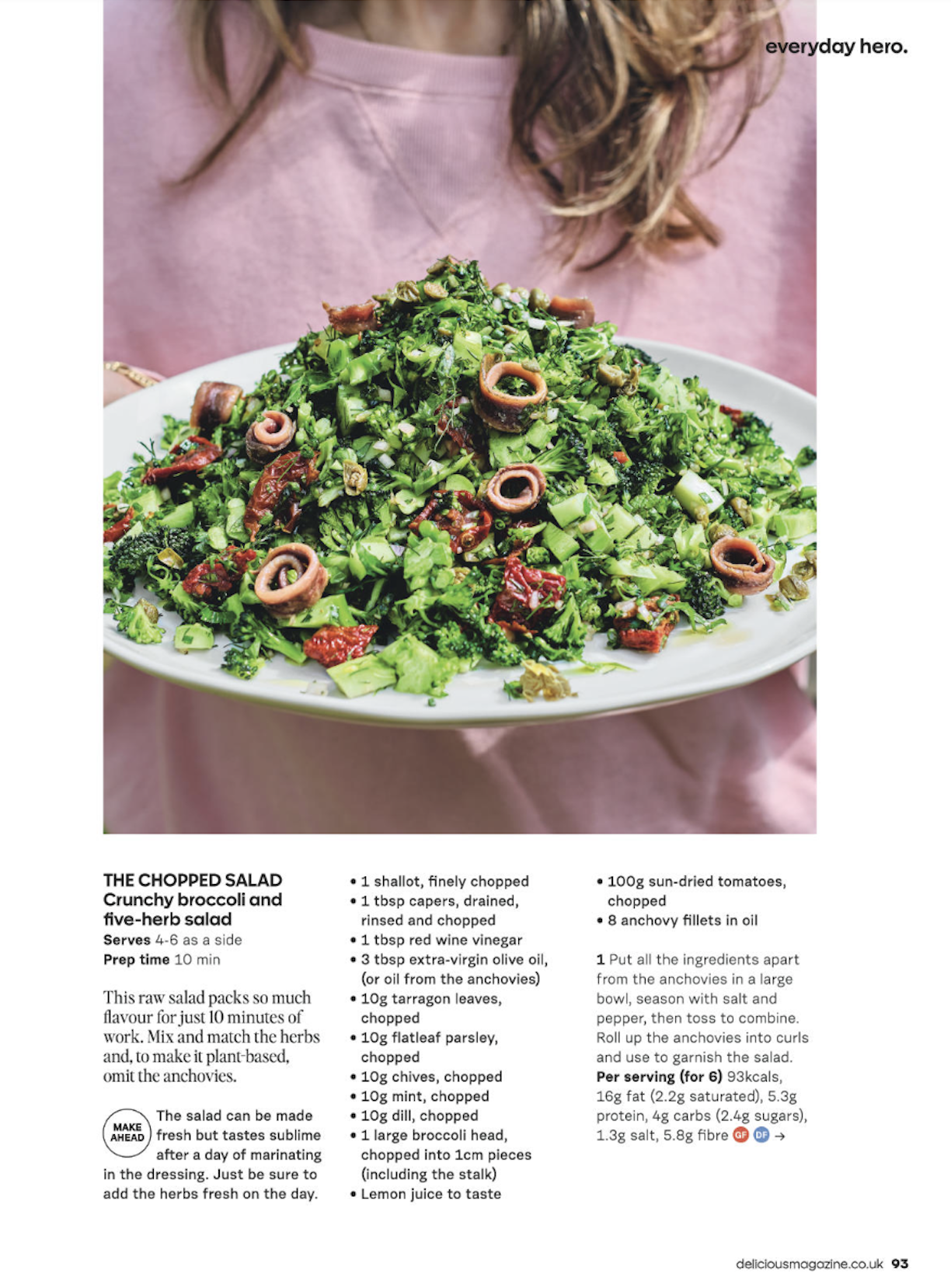 A person holding a large white plate with a colorful chopped salad consisting of broccoli, sun-dried tomatoes, anchovy fillets, and herbs. The person is wearing a pink shirt, and the background contains a small text "everyday hero."