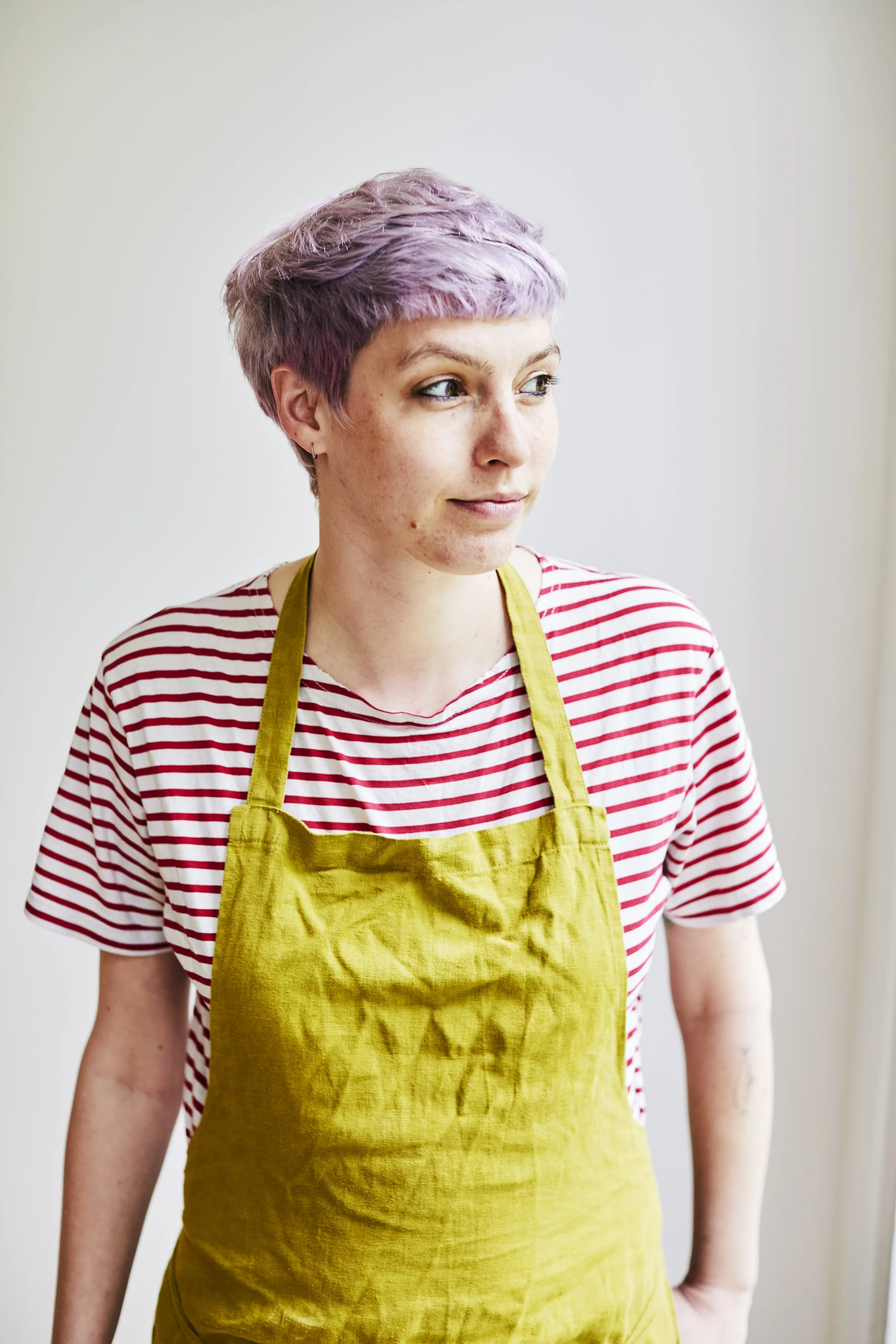 Young person with short purple hair wearing a chartreuse coloured apron and a red and white striped T-shirt looking to the side.