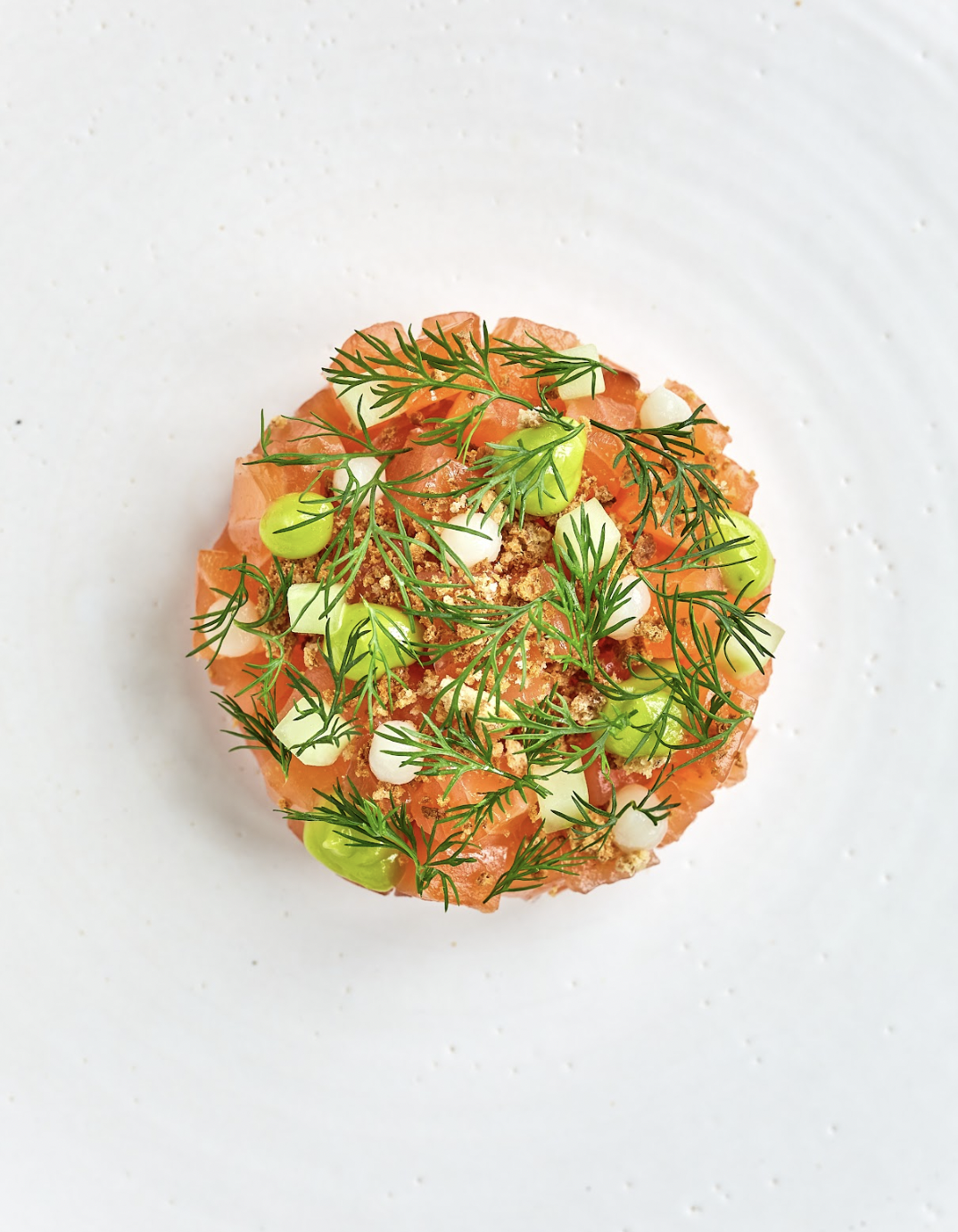 A round, colorful dish with diced salmon, green herbs, and edible flowers garnished on a white plate.