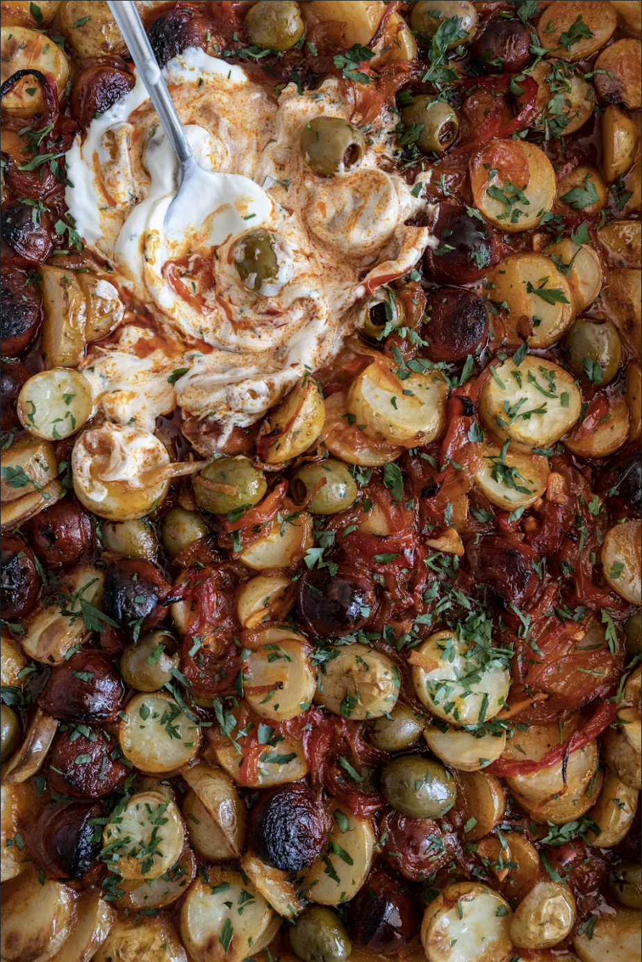Close-up of roasted potatoes and vegetables topped with chopped fresh herbs, with a spoonful of sour cream and some spices on top.