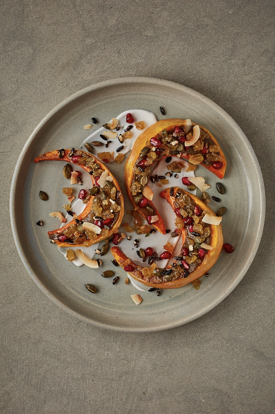 Roasted squash slices topped with a mixture of seeds, dried fruits, and coconut flakes, served on a plate with a drizzle of yogurt or sauce.