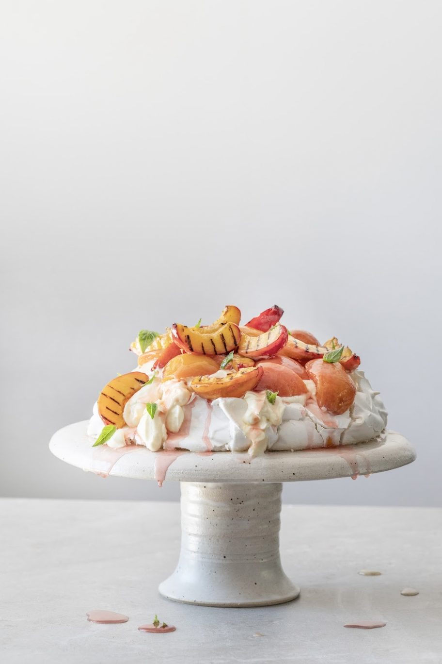 A Pavlova dessert topped with sliced peaches, strawberries, apricots, chopped nuts, and small green leaves on a white pedestal