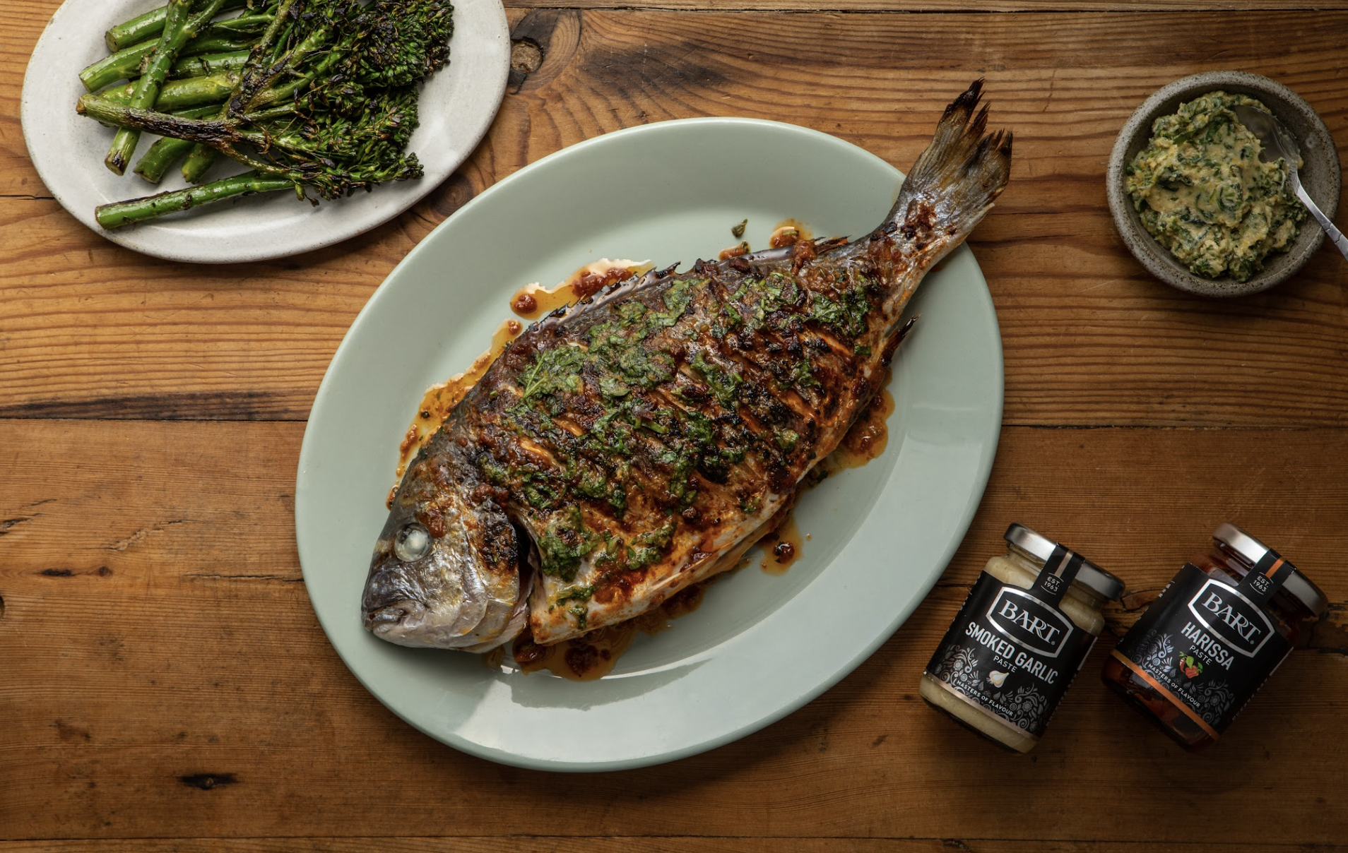 A grilled whole fish garnished with herbs on a plate, served with roasted green asparagus on a side plate, a small bowl of herb sauce, and two jars labeled smoked garlic and harissa on a wooden table.